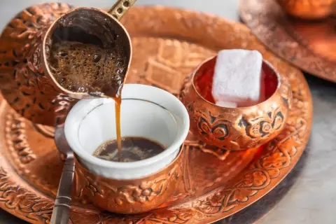 Frisch aufgebrühter, bester türkischer Kaffee fließt aus einer Kupferkanne in eine kleine Tasse. Daneben liegen süße Loukoum-Würfel.